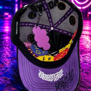May include: A purple and black trucker hat with a mesh back, featuring a purple cloud-shaped tag inside. The interior lining displays colorful circular patches with the text DH and SAD BOYZ. The brim has white embroidered text and black script.