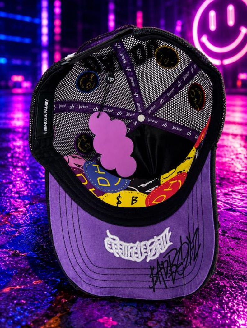 May include: A purple and black trucker hat with a mesh back, featuring a purple cloud-shaped tag inside. The interior lining displays colorful circular patches with the text DH and SAD BOYZ. The brim has white embroidered text and black script.