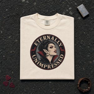 May include: A cream-colored t-shirt featuring a circular graphic of a vampire with dark hair, red eyeshadow, and fangs. The text "ETERNALLY UNIMPRESSED" is printed in a circular border around the illustration, which also includes small red bats.