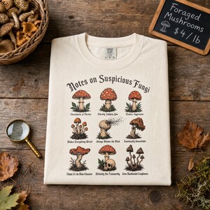 May include: A cream-colored t-shirt featuring a graphic titled Notes on Suspicious Fungi. The design displays nine distinct mushroom illustrations, each paired with a humorous caption such as Overshares at Parties and Emotionally Unavailable.