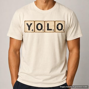 May include: A cream-colored short-sleeve t-shirt featuring a graphic print of four square tiles resembling a word game. The tiles display the letters Y, O, L, O in black, each with a small numerical value in the corner, arranged horizontally.