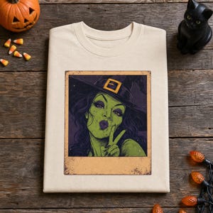 May include: A cream-colored t-shirt featuring a rectangular graphic print of a green-skinned witch wearing a dark purple pointed hat with a yellow buckle. The witch has dark hair, purple lipstick, and is making a peace sign with her fingers.