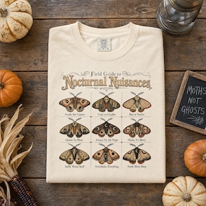 Può includere: Una t-shirt color crema con una grafica intitolata Field Guide to Nocturnal Nuisances. Il design mostra nove falene illustrate con motivi unici ed etichette come Avoids Eye Contact, Cries at Candles e Bad at Parties.