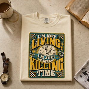 May include: A cream-colored t-shirt featuring a graphic design with a clock face and the text I'M NOT LIVING, I'M JUST KILLING TIME. The design uses yellow, blue, and black colors with a checkered border and star accents on a short-sleeve shirt.