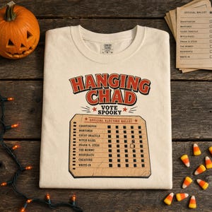 May include: A cream-coloured t-shirt featuring the text HANGING CHAD VOTE SPOOKY in red and black. Below is a graphic of an official election ballot with names like Ghostington and Count Dracula, with a checkmark next to Witch Hazel.