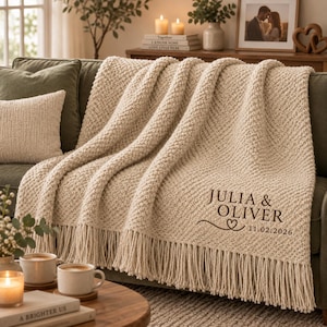 May include: A cream-colored knitted throw blanket with a textured pattern and long fringe along the bottom edge. The blanket features dark brown embroidered text that reads JULIA & OLIVER, a small heart icon, and the date 11.02.2026.