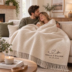 May include: A cream-colored, textured knit throw blanket featuring a fringe trim along the bottom edge. The blanket is personalized with a black printed design consisting of a heart icon, the letters A and M, the names Amelia & William, and 05.28.2026.