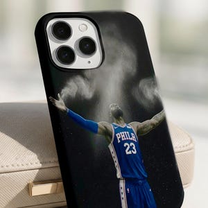 Puede incluir: Una funda de smartphone negra con la imagen de un jugador de baloncesto con camiseta azul y el texto PHILA 23, brazos extendidos con nubes de polvo blanco sobre un fondo oscuro y una abertura para la triple lente de la cámara.