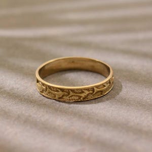 Puede incluir: Un anillo de metal en tono dorado con un diseño de banda continua que presenta un patrón repetitivo de pequeñas siluetas de peces estilizadas en relieve en la superficie exterior, sobre un fondo texturizado de acabado mate.
