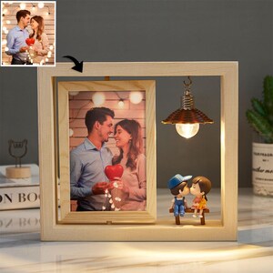 May include: A rectangular light-colored wooden frame containing a vertical photograph of a couple holding a red heart. To the right, a small hanging lamp illuminates two miniature figurines of a boy and girl sitting on a wooden bench.