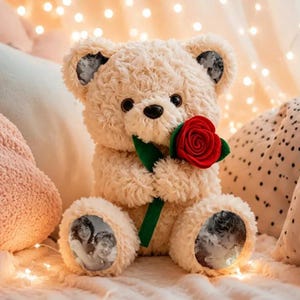 Puede incluir: Un oso de peluche beige con pelaje rizado, sosteniendo una rosa roja con hojas verdes. El oso presenta impresiones fotográficas circulares en blanco y negro en las orejas y los pies, mostrando retratos de dos personas en un abrazo cercano.
