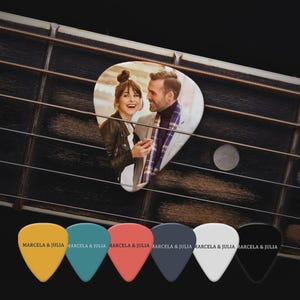 May include: A guitar plectrum featuring a printed photograph of a smiling couple, positioned on guitar strings. Below are six solid-coloured guitar plectrums in yellow, teal, coral, dark grey, white, and black, each printed with the text MARCELA & JULIA.