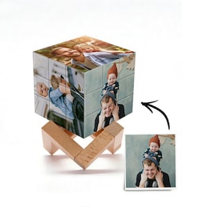 May include: A multi-faceted photo cube puzzle featuring printed images of people on its surfaces, supported by a light-coloured wooden stand. The cube displays various family photographs, and a separate square print of one image is shown nearby.