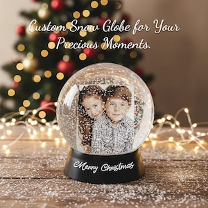 May include: A clear glass snow globe featuring a rectangular photograph of two children inside. The black base is inscribed with the white text Merry Christmas. The text Custom Snow Globe for Your Precious Moments is visible above the globe.