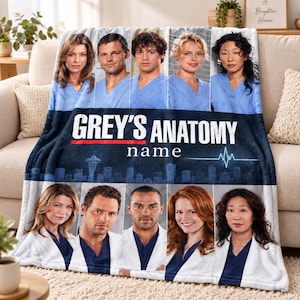 May include: A soft fleece blanket featuring a Grey's Anatomy graphic with the text "GREY'S ANATOMY" and "name" above a Seattle skyline silhouette. The design includes two rows of portraits of the show's characters wearing blue scrubs and white coats.