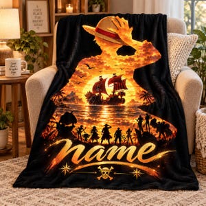 Peut inclure: Une couverture en polaire noire ornée d'un coucher de soleil orange et jaune vif avec un navire pirate, des silhouettes de personnages et un grand chapeau de paille. Le mot name est imprimé en écriture cursive au-dessus d'un crâne et d'os.
