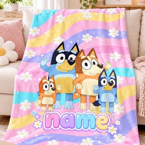 May include: A soft fleece blanket featuring a colourful wavy background of pink, yellow, and blue stripes with white daisy flowers. The design includes four cartoon dog characters and the word name in a large, rounded, light blue font at the bottom.