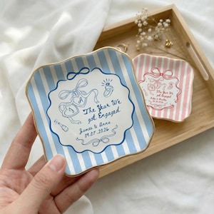 May include: A square ceramic trinket dish with wavy edges, featuring light blue and white vertical stripes. The center displays a blue graphic of a bow, a diamond ring, a champagne bottle, and the text The Year We got Engaged, James & Anna, 09.07.2026.