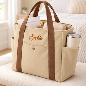 May include: A beige canvas tote bag with brown fabric handles and trim. The front features the name Sophia embroidered in brown script. The bag includes a front pocket with a gold snap button and side pockets containing a white tumbler.