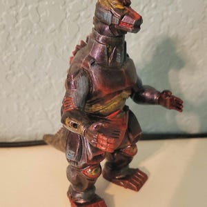 May include: A mechanical monster figurine featuring a dark metallic finish with bronze and copper highlights. The creature has a robotic appearance with segmented armor plating, a pointed snout, and clawed feet, standing in an upright posture.