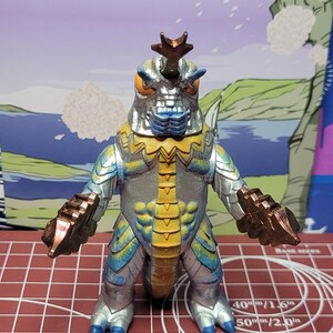 May include: A monster figurine with a silver body, yellow chest, and metallic bronze claws and head horn. The creature has orange eyes and blue accents on its feet and face. It stands on a red cutting mat with white grid lines and measurement text.