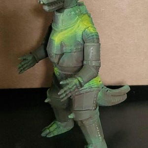 May include: A green and brown mechanical monster figurine with a reptilian shape, featuring a textured metallic-looking body, segmented limbs, a long tail, and a distinct yellow eye area. The figure stands in a bipedal pose with arms extended.