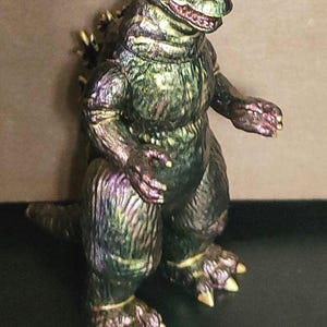 May include: A standing monster figurine with a textured, iridescent finish that shifts between shades of dark green, purple, and bronze. The creature features a reptilian head, sharp claws on its hands and feet, and prominent dorsal fins on its back.