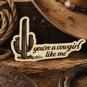 May include: A sticker with a cactus illustration and the text "you're a cowgirl like me." The sticker is cream-colored with black details. A brown cowboy hat, rope, and horseshoe are in the background.