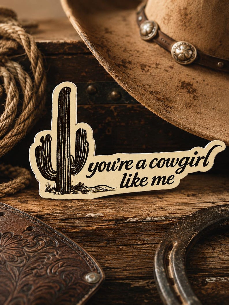 May include: A sticker with a cactus illustration and the text "you're a cowgirl like me." The sticker is cream-colored with black details. A brown cowboy hat, rope, and horseshoe are in the background.