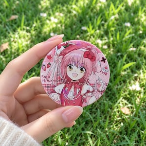 Puede incluir: Un botón circular con alfiler que muestra un personaje de anime de cabello rosa con un lazo, rodeado de ilustraciones de fresas y pasteles. El texto Amu Hinamori está impreso a la izquierda. El botón tiene un acabado brillante y tonos rosas.