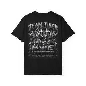 Puede incluir: Una camiseta negra de manga corta con un gráfico de un tigre rugiendo con llaves inglesas cruzadas. El texto dice TEAM TIGER, RWE, MAINTENENCE AND OPERATIONS, y I CAN DO ALL THINGS THROUGH CHRIST WHO STRENGTHENS ME. PHILIPPIANS 4:13.