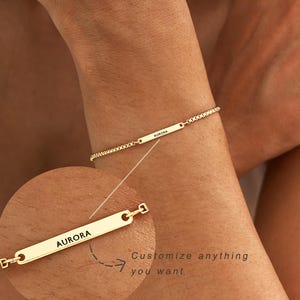 May include: A gold-toned chain bracelet featuring a thin, rectangular metal bar engraved with the name AURORA. A circular inset shows a magnified view of the bar with the text AURORA and the additional text Customize anything you want.