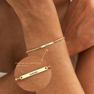 Peut inclure: Un bracelet à chaîne doré doté d'une fine barre métallique rectangulaire gravée du nom AURORA. Un encart circulaire montre une vue agrandie de la barre avec le texte Customize anything you want, soulignant l'option de personnalisation.