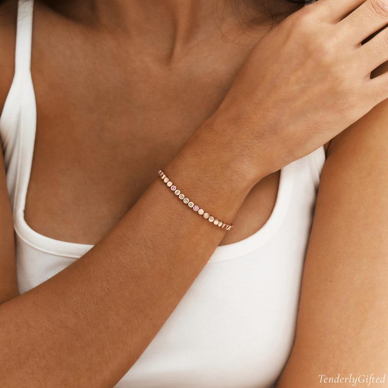 May include: A rose gold-toned bracelet featuring a series of small, circular links. Several of these links are personalized with individual letters that spell out the name EMMA, with the letter A highlighted in a distinct pink circular setting.
