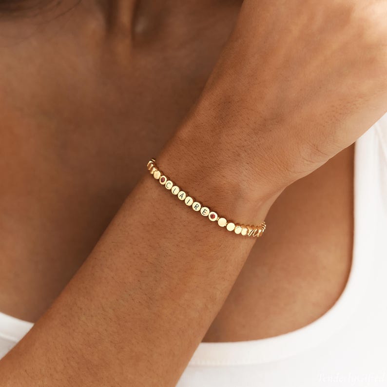 May include: A gold-toned bracelet featuring a series of small, circular, flat beads. Several of these beads are engraved with the letters spelling CLAIRE, while two small, round, reddish-brown stones are positioned at the ends of the name sequence.