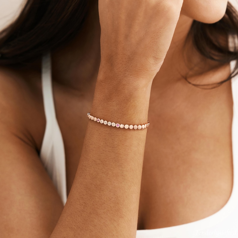 May include: A rose gold-toned bracelet featuring a series of small, circular, metallic discs. Several of these discs are engraved with the letters E, M, M, and A, while one disc displays a small, circular pink accent stone in the center.