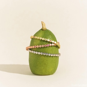 May include: Three beaded bracelets in gold, rose gold, and silver tones wrapped around a green fig. The gold bracelet features the name CLAIRE, the rose gold bracelet features the name Emma, and the silver bracelet features the numbers 0712.