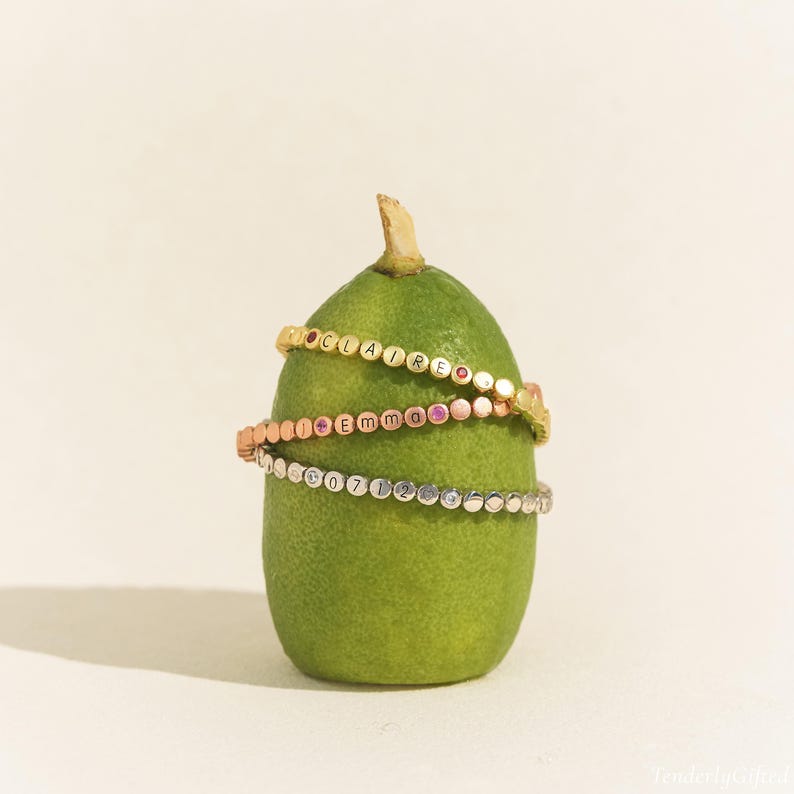 May include: Three beaded bracelets in gold, rose gold, and silver tones wrapped around a green fig. The gold bracelet features the name CLAIRE, the rose gold bracelet features the name Emma, and the silver bracelet features the numbers 0712.