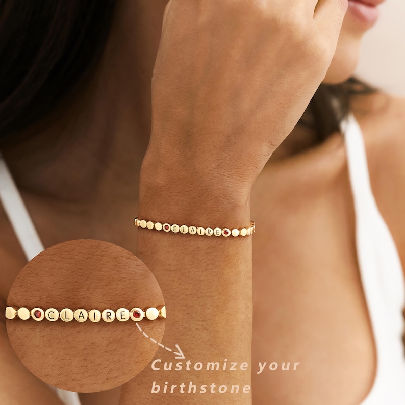 May include: A gold-toned bracelet featuring a series of small, circular beads. The beads spell out the name CLAIRE in black capital letters, interspersed with two small, round, red gemstones. The bracelet is shown on a wrist and in a circular inset.