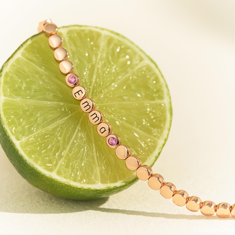 May include: A rose gold-toned beaded bracelet featuring small round beads, two pink circular gemstones, and four beads engraved with the letters E, M, M, and A, arranged in a sequence that spells out the name Emma along the length of the chain.