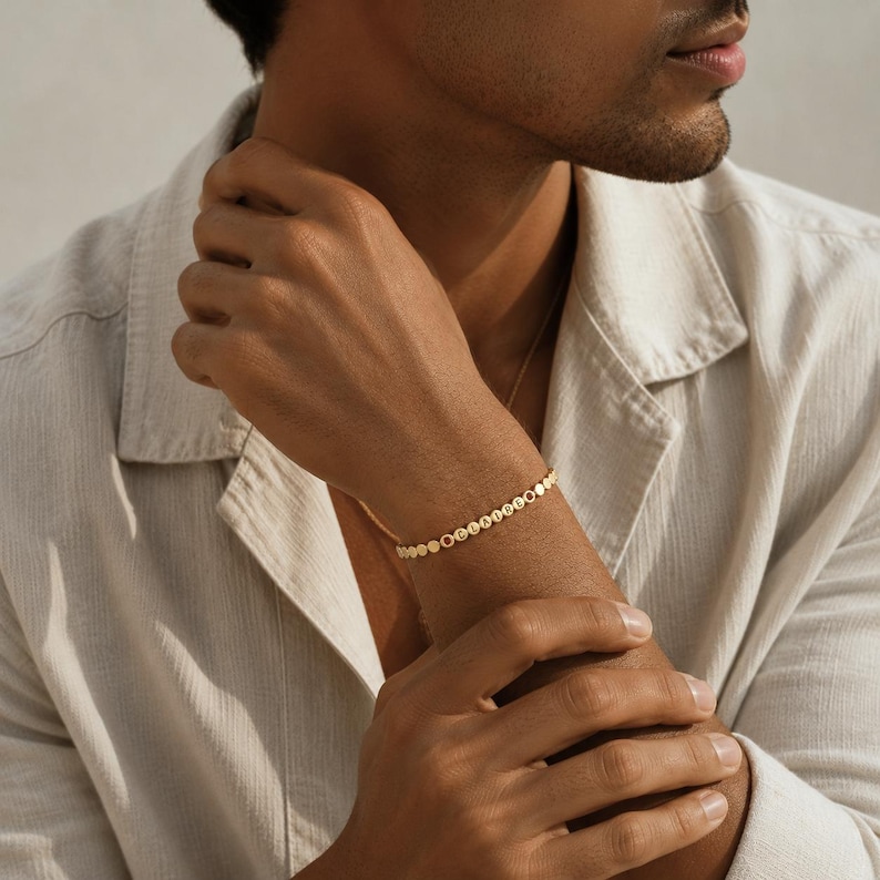May include: A gold-toned bracelet featuring a series of small, circular, flat discs linked together. Each disc is embossed with a single letter, collectively spelling out the name CLAIRE in a horizontal sequence around the wrist.