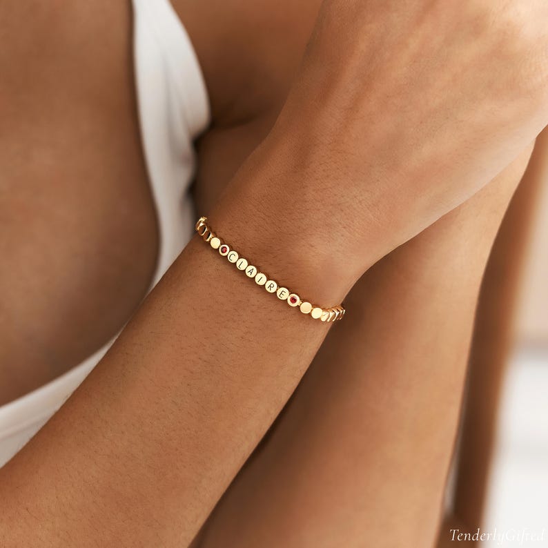 May include: A gold-toned beaded bracelet featuring small, round, metallic beads. The bracelet includes letter beads spelling out the name CLAIRE, interspersed with two small, circular, red-colored accent beads positioned within the sequence.