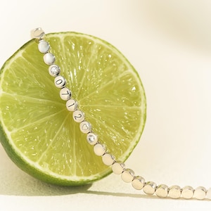 May include: A silver-toned bracelet composed of small, circular metal discs linked together. Several discs feature engraved numbers and a heart symbol, while others are set with small, clear, round stones, resting against a sliced green lime.