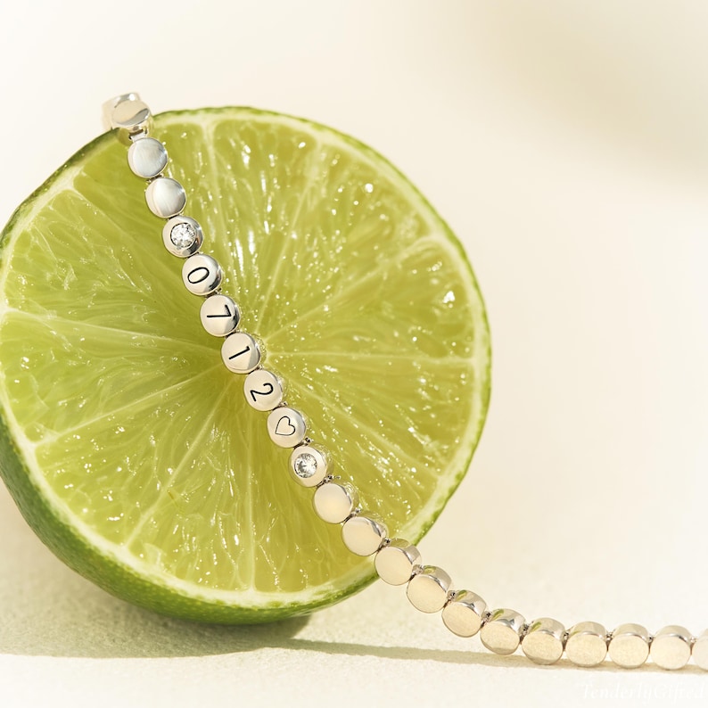 May include: A silver-toned bracelet composed of small, circular metal discs linked together. Several discs feature engraved numbers and a heart symbol, while others are set with small, clear, round stones, resting against a sliced green lime.