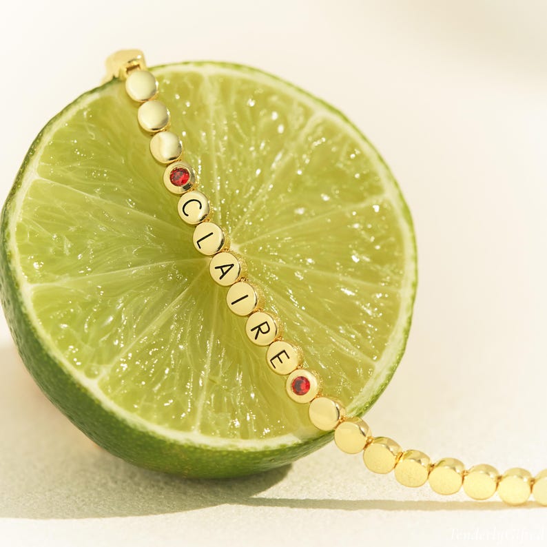 May include: A gold-toned bracelet featuring small, round, flat beads. The beads spell out the name CLAIRE in black capital letters, interspersed with two small, circular, vibrant red stones. The bracelet is draped across a sliced green lime.