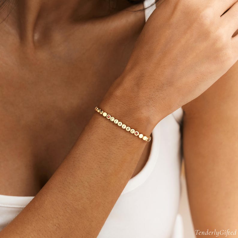 May include: A gold-toned beaded bracelet featuring small, round, metallic discs. The bracelet includes a series of circular beads with the letters C, L, A, I, R, E embossed in black, interspersed with small, plain gold-colored beads along the chain.