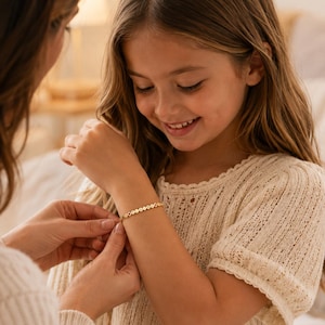 May include: A gold-toned bracelet featuring a series of small, circular, flat charms linked together. Each charm is engraved with a single letter, creating a personalized name or word sequence that wraps around the wrist in a delicate, minimalist style.
