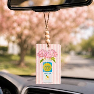 May include: A rectangular car air freshener hanging from a rearview mirror by a twine string with three wooden beads. The freshener features a pink and white striped background with a graphic of a High Noon pineapple vodka and soda can.