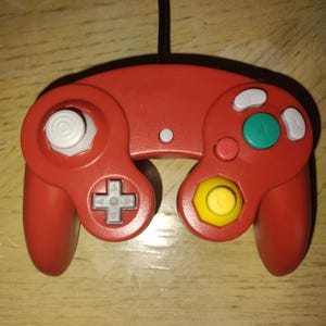 May include: A red wired video game controller featuring a white analog stick on the left, a yellow C-stick on the right, a gray directional pad, a large green A button, a red B button, and white X and Y buttons, with a black cable extending upward.