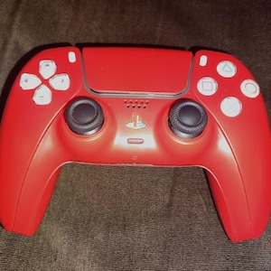 Op de afbeelding: Een rode gamecontroller met witte knoppen en zwarte joysticks. De controller heeft een PlayStation-logo en een centraal touchpad. De knoppen omvatten een D-pad, cirkel, vierkant, driehoek en X.